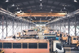A large indoor facility filled with industrial vehicles and machinery. The space is structured with metal beams and bright overhead lighting. There are multiple heavy-duty trucks and construction equipment lined up, suggesting a maintenance or storage area. The setting appears organized with cranes and equipment systematically arranged.