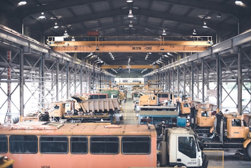A large indoor facility filled with industrial vehicles and machinery. The space is structured with metal beams and bright overhead lighting. There are multiple heavy-duty trucks and construction equipment lined up, suggesting a maintenance or storage area. The setting appears organized with cranes and equipment systematically arranged.