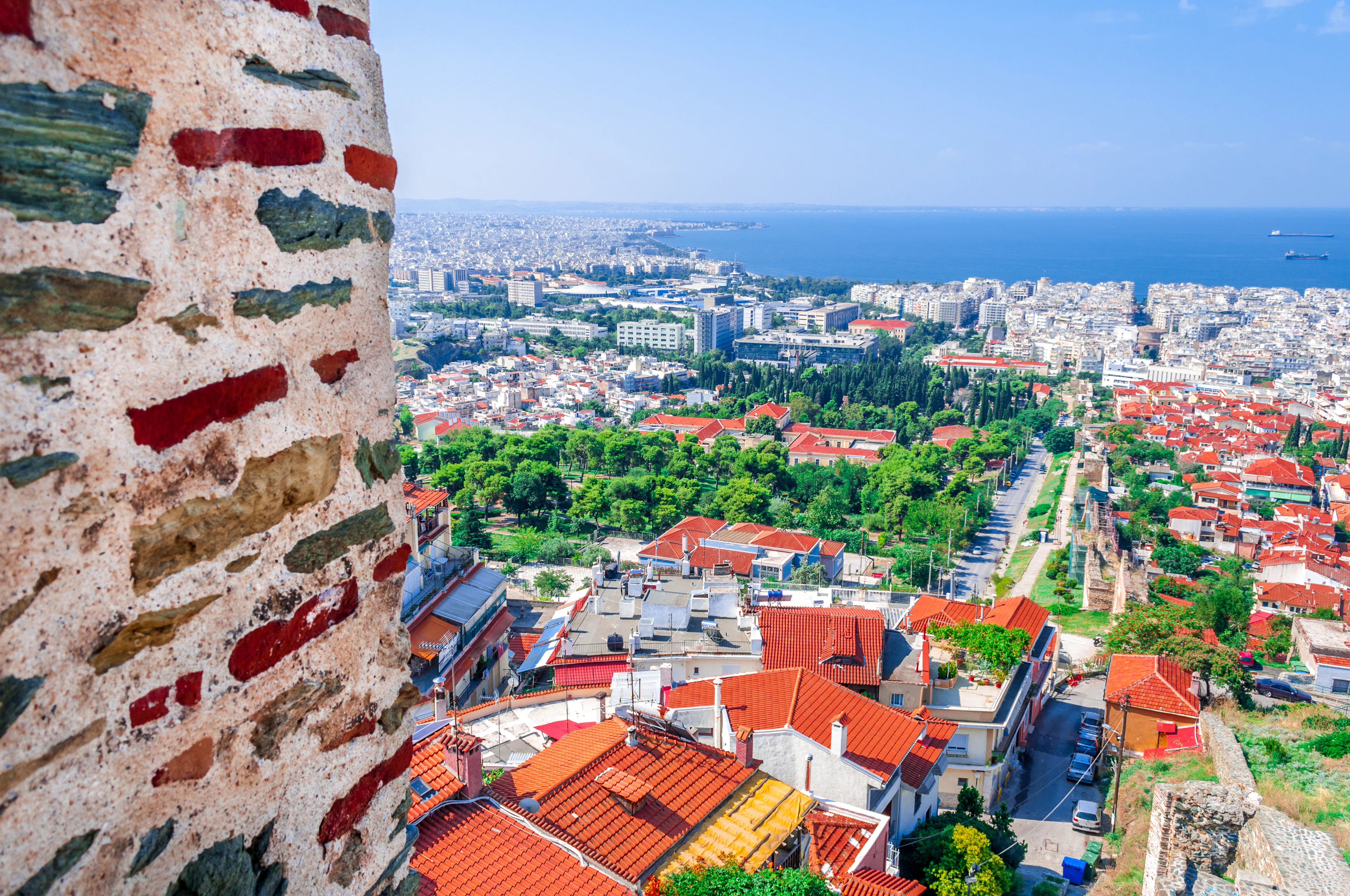 Thessaloniki seen from the Byzantine walls of the upper town, red-tiled roofs and white apartment blocks running downhill to the blue Thermaic Gulf, a band of ancient brick-and-stone rampart in the left foreground