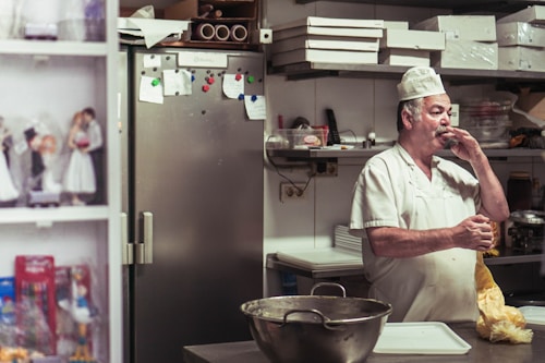 A man wearing a white chef's uniform and a cap is standing in a kitchen. He appears to be eating or tasting something while holding a piece of food in his hand. In the background, there are kitchen items, including a large steel bowl on the counter, several sheets of paper attached to a stainless steel refrigerator, and boxes stacked on top of shelves. To the left, there are figurines on a shelf.