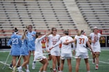 A group of female lacrosse players on a field, with some wearing blue uniforms and others in white. They appear to be in a huddle or discussion, holding lacrosse sticks. The background shows empty bleachers and a green field.