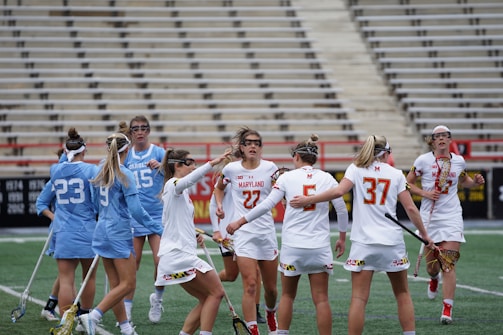 A group of female lacrosse players on a field, with some wearing blue uniforms and others in white. They appear to be in a huddle or discussion, holding lacrosse sticks. The background shows empty bleachers and a green field.
