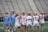 A group of female lacrosse players on a field, with some wearing blue uniforms and others in white. They appear to be in a huddle or discussion, holding lacrosse sticks. The background shows empty bleachers and a green field.