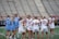 A group of female lacrosse players on a field, with some wearing blue uniforms and others in white. They appear to be in a huddle or discussion, holding lacrosse sticks. The background shows empty bleachers and a green field.