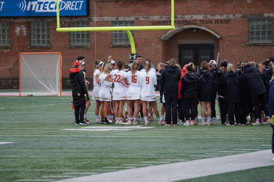 A group of athletes wearing sports uniforms huddles on a grassy field, likely preparing or strategizing for a game. The players are surrounded by additional team members or coaches wearing black jackets. A goalpost and net are visible in the background, along with a brick building.
