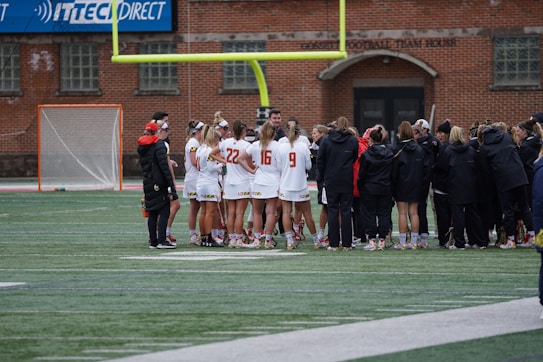 A group of athletes wearing sports uniforms huddles on a grassy field, likely preparing or strategizing for a game. The players are surrounded by additional team members or coaches wearing black jackets. A goalpost and net are visible in the background, along with a brick building.