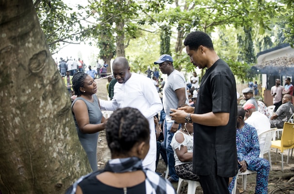 A group of people gathered outdoors in a lush, green environment. Some individuals are engaged in conversation while others are seated. Various people are dressed in casual or traditional attire, and the setting appears to be informal and social.