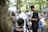 A group of people gathered outdoors in a lush, green environment. Some individuals are engaged in conversation while others are seated. Various people are dressed in casual or traditional attire, and the setting appears to be informal and social.