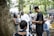 A group of people gathered outdoors in a lush, green environment. Some individuals are engaged in conversation while others are seated. Various people are dressed in casual or traditional attire, and the setting appears to be informal and social.
