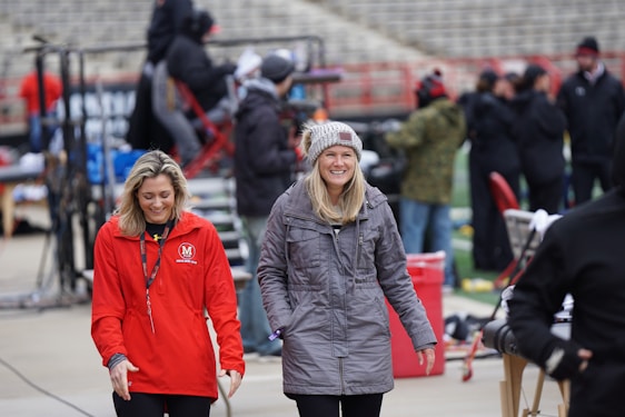 A stylish woman bundled in warm clothing watching a youth sports game outdoors.