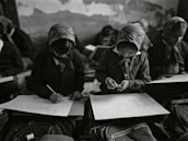 In a classroom setting, several students wearing headscarves are seated closely together. They are focused on drawing sketches on large sheets of paper, with bags and other materials visible around them. The classroom has an intimate and focused atmosphere.
