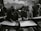 In a classroom setting, several students wearing headscarves are seated closely together. They are focused on drawing sketches on large sheets of paper, with bags and other materials visible around them. The classroom has an intimate and focused atmosphere.