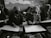 In a classroom setting, several students wearing headscarves are seated closely together. They are focused on drawing sketches on large sheets of paper, with bags and other materials visible around them. The classroom has an intimate and focused atmosphere.