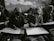 In a classroom setting, several students wearing headscarves are seated closely together. They are focused on drawing sketches on large sheets of paper, with bags and other materials visible around them. The classroom has an intimate and focused atmosphere.