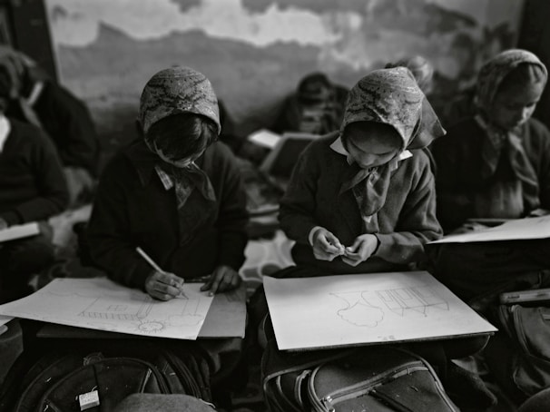 In a classroom setting, several students wearing headscarves are seated closely together. They are focused on drawing sketches on large sheets of paper, with bags and other materials visible around them. The classroom has an intimate and focused atmosphere.