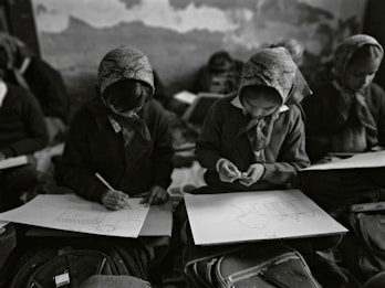 In a classroom setting, several students wearing headscarves are seated closely together. They are focused on drawing sketches on large sheets of paper, with bags and other materials visible around them. The classroom has an intimate and focused atmosphere.