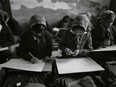 In a classroom setting, several students wearing headscarves are seated closely together. They are focused on drawing sketches on large sheets of paper, with bags and other materials visible around them. The classroom has an intimate and focused atmosphere.