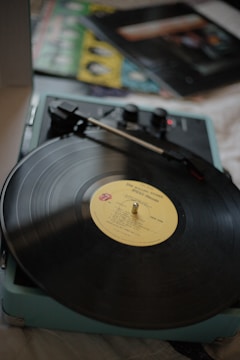 A vintage record player is playing a vinyl disk labeled with album information. The record player has a light blue casing with a spindle and a tonearm. In the background, there are partially visible album covers with vibrant colors.