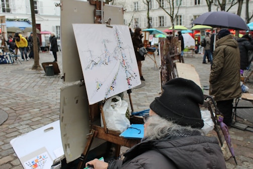A street scene in an outdoor art market or fair. An artist sits in the foreground, working on an abstract painting, with a palette and other art supplies nearby. Several other easels with canvases are visible, indicating more artists at work. People are walking around, some holding umbrellas, suggesting a possibly rainy day. The background includes townhouses with trees lining the area.