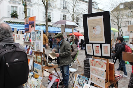 A street art market features artists displaying various paintings on easels and stands. Several people, dressed in winter attire, browse the artwork amidst a lively, bustling environment with vibrant umbrellas providing shelter.