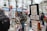 A street art market features artists displaying various paintings on easels and stands. Several people, dressed in winter attire, browse the artwork amidst a lively, bustling environment with vibrant umbrellas providing shelter.