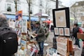 A street art market features artists displaying various paintings on easels and stands. Several people, dressed in winter attire, browse the artwork amidst a lively, bustling environment with vibrant umbrellas providing shelter.
