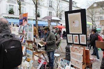 A street art market features artists displaying various paintings on easels and stands. Several people, dressed in winter attire, browse the artwork amidst a lively, bustling environment with vibrant umbrellas providing shelter.