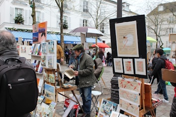 A street art market features artists displaying various paintings on easels and stands. Several people, dressed in winter attire, browse the artwork amidst a lively, bustling environment with vibrant umbrellas providing shelter.