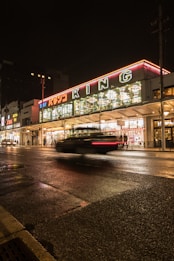 A nighttime street scene features a brightly illuminated building with the word 'KING' in large letters. The foreground shows a motion-blurred car driving past on the wet road, reflecting the building's lights. The building has multiple signs with colorful neon lights, and there are people walking and standing along the sidewalk.