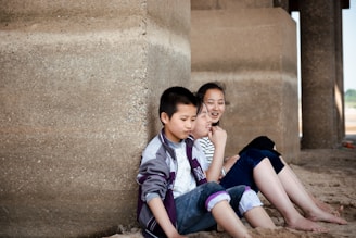 Three children are sitting on the sand, leaning against a large concrete structure. They appear relaxed and are smiling, suggesting they are enjoying their time together.
