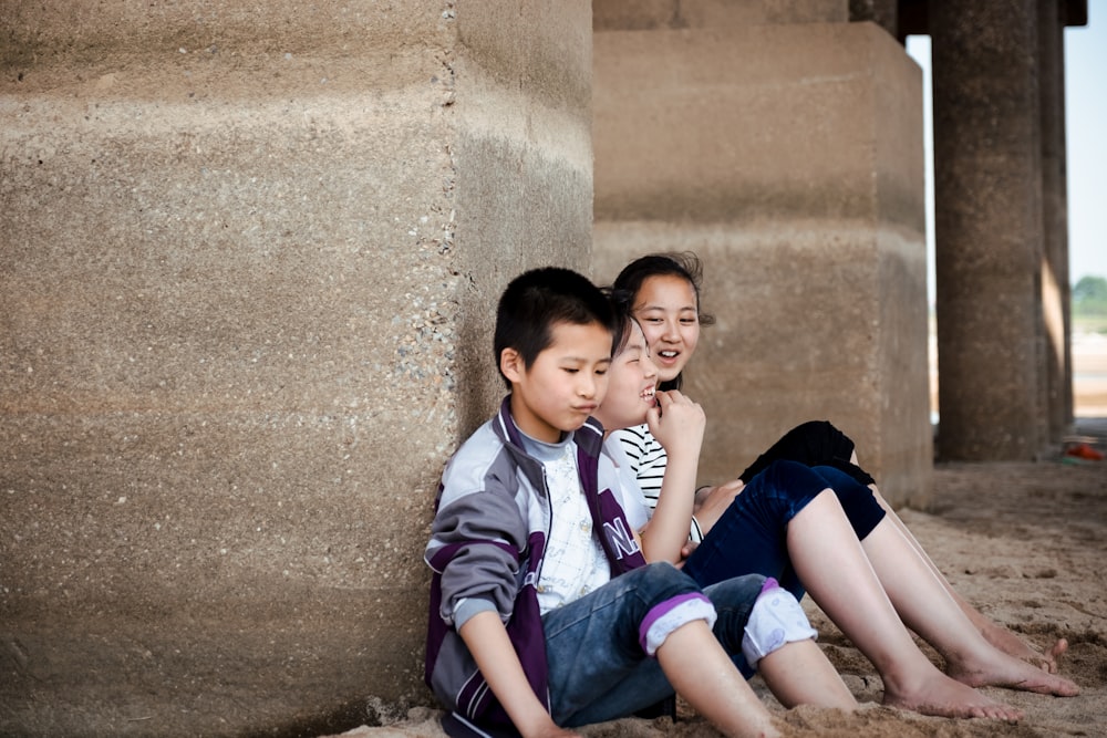 three kids sitting on sand and leaning on concrete wall during daytime