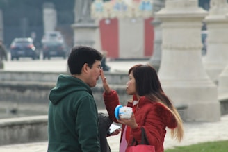 A woman is applying cream to a man's face in an outdoor setting. They appear to be standing near a railing and large stone pillars. The woman is holding a container of cream in one hand and a red bag hangs on her arm. In the background, there are blurred cars and structures.