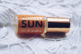 A small perfume bottle labeled 'Summer Sun' lies on a textured white lace fabric. The bottle features a golden cap and an amber liquid inside.