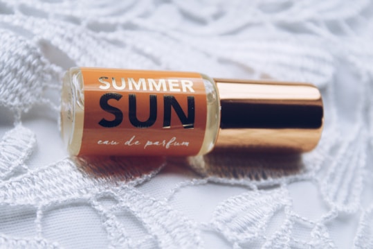 A small perfume bottle labeled 'Summer Sun' lies on a textured white lace fabric. The bottle features a golden cap and an amber liquid inside.