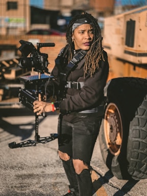 A person wearing a black outfit and a bandana operates a steadicam rig outdoors, next to a large vehicle with visible wheels. The setting appears industrial, with scattered shadows and a bright sunlight casting contrast across the scene.