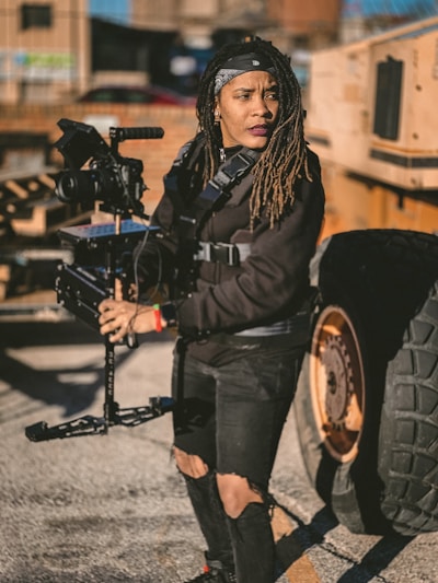 A person wearing a black outfit and a bandana operates a steadicam rig outdoors, next to a large vehicle with visible wheels. The setting appears industrial, with scattered shadows and a bright sunlight casting contrast across the scene.