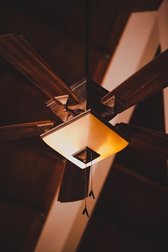 A wooden ceiling fan with a square-shaped light fixture, featuring four blades extending outwards. The light has a warm glow, with pull chains hanging down. The ceiling appears to have a wooden texture, adding to the rustic ambiance.
