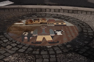 A mosaic embedded in cobblestone depicts a rustic scene, featuring a farmer with a large moustache and wide-brimmed hat, wearing boots and a vest. Beside him are cows, and behind, a line of quaint houses with thatched roofs is visible.