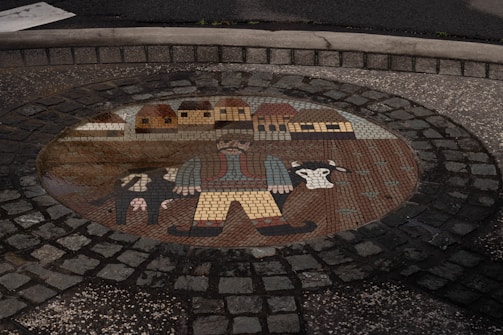 A mosaic embedded in cobblestone depicts a rustic scene, featuring a farmer with a large moustache and wide-brimmed hat, wearing boots and a vest. Beside him are cows, and behind, a line of quaint houses with thatched roofs is visible.