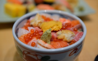 A close-up view of a bowl containing a colorful assortment of seafood, including salmon roe, sea urchin, and various cuts of fish. The bowl features a decorative floral design and is placed against a blurred background, highlighting the texture and colors of the dish.