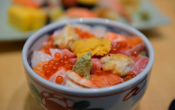 A close-up view of a bowl containing a colorful assortment of seafood, including salmon roe, sea urchin, and various cuts of fish. The bowl features a decorative floral design and is placed against a blurred background, highlighting the texture and colors of the dish.