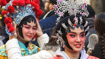 Two individuals in traditional, elaborate Chinese opera costumes with intricate headpieces adorned with silver ornaments and red embellishments. Their makeup is vibrant, with dramatic eye designs and rosy cheeks, suggesting a performance setting. The background includes other blurred figures, indicating a crowded or public gathering.