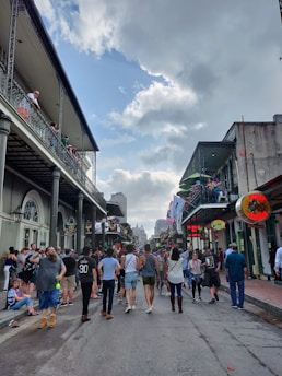 A busy street scene with people walking and socializing. There are buildings on either side featuring balconies, with some individuals watching from above. The street is lively with a diverse crowd, including couples and groups, creating a festive atmosphere. Flags and signs add color and vibrancy to the scene, while the sky is partly cloudy.