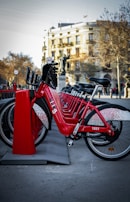 A line of red rental bicycles is parked in an organized row at a docking station. The bicycles are bright and prominently displayed in the foreground. Behind them, there's a cityscape that includes a building with classic architecture and leafless trees, indicating a likely autumn or winter setting.