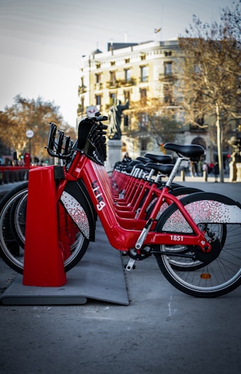 A line of red rental bicycles is parked in an organized row at a docking station. The bicycles are bright and prominently displayed in the foreground. Behind them, there's a cityscape that includes a building with classic architecture and leafless trees, indicating a likely autumn or winter setting.