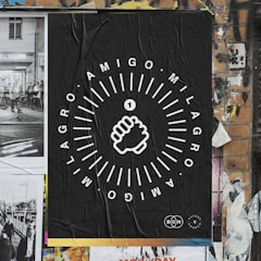 A poster on a brick wall featuring a black background with white graphic design. The central image is a stylized fist surrounded by circular textual elements reading 'AMIGO MILAGRO'. The wall includes torn and overlapping posters, contributing to a textured, urban street art aesthetic.