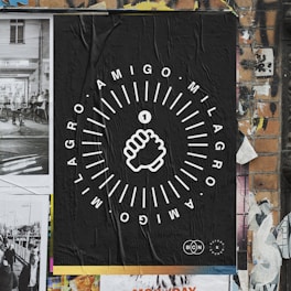 A poster on a brick wall featuring a black background with white graphic design. The central image is a stylized fist surrounded by circular textual elements reading 'AMIGO MILAGRO'. The wall includes torn and overlapping posters, contributing to a textured, urban street art aesthetic.