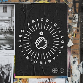 A poster on a brick wall featuring a black background with white graphic design. The central image is a stylized fist surrounded by circular textual elements reading 'AMIGO MILAGRO'. The wall includes torn and overlapping posters, contributing to a textured, urban street art aesthetic.