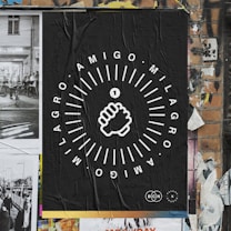 A poster on a brick wall featuring a black background with white graphic design. The central image is a stylized fist surrounded by circular textual elements reading 'AMIGO MILAGRO'. The wall includes torn and overlapping posters, contributing to a textured, urban street art aesthetic.