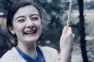 A young person is laughing joyfully while holding onto a rope. The background is slightly blurred with hints of greenery and a wooden structure.